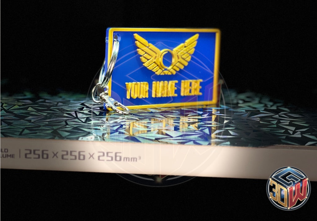 3D-printed, blue and gold name tag with a space-themed design, featuring a winged emblem and customizable text area labeled 'Your Name Here.' The tag is rectangular, lightweight, and attached to a keyring, sitting on a 3D printer bed.