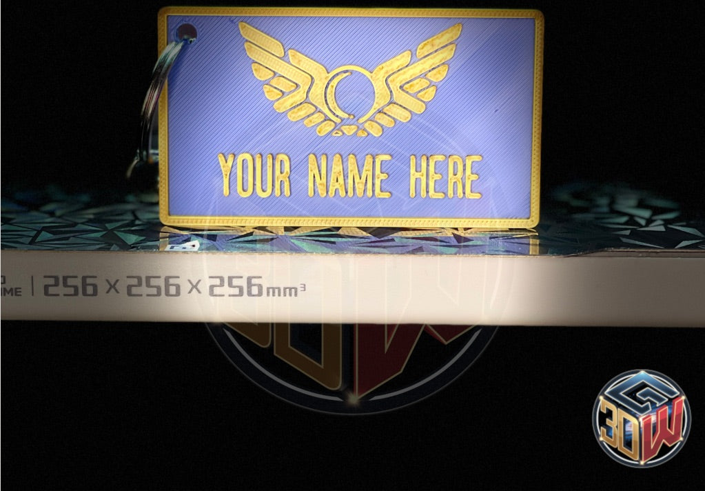 3D-printed, blue and gold name tag with a space-themed design, featuring a winged emblem and customizable text area labeled 'Your Name Here.' The tag is rectangular, lightweight, and attached to a keyring, sitting on a 3D printer bed facing forward.